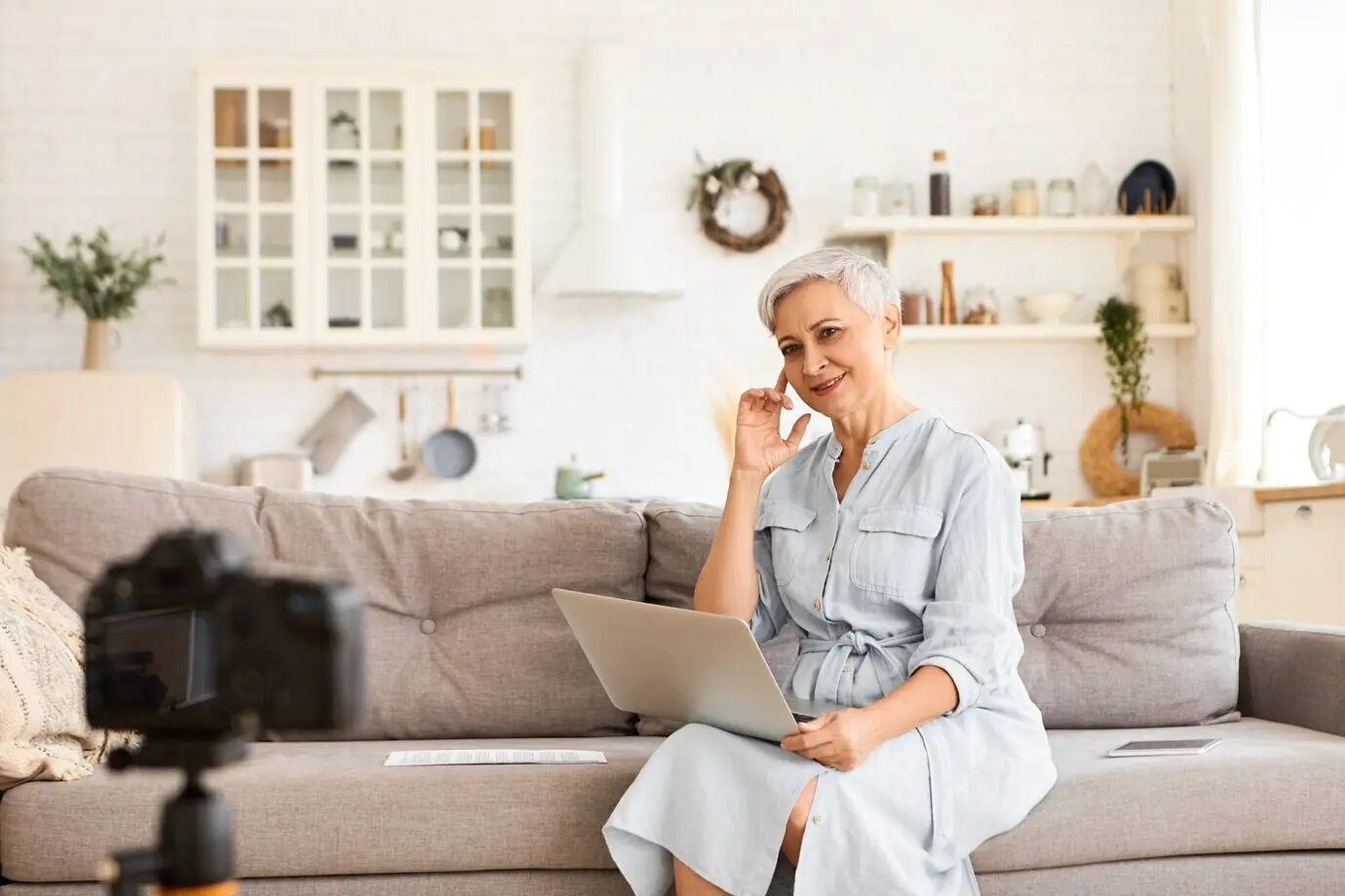 Konzept zu Menschen, Alter, Reife und moderner Technologie: Innenaufnahme einer stilvollen, kurzhaarigen Bloggerin, die im Wohnzimmer auf dem Sofa ein Video aufnimmt, auf dem Laptop tippt und den Blick fest auf das Stativ richtet.